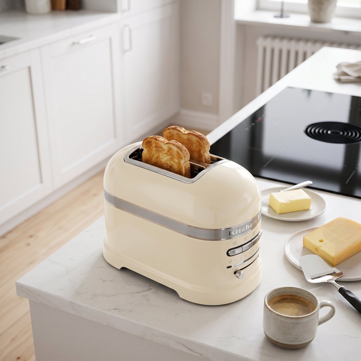 Krämvit KitchenAid Artisan med rostat bröd på en vit bänk bredvid smör, ost och osthyvel i ett svenskt kök med induktionshäll