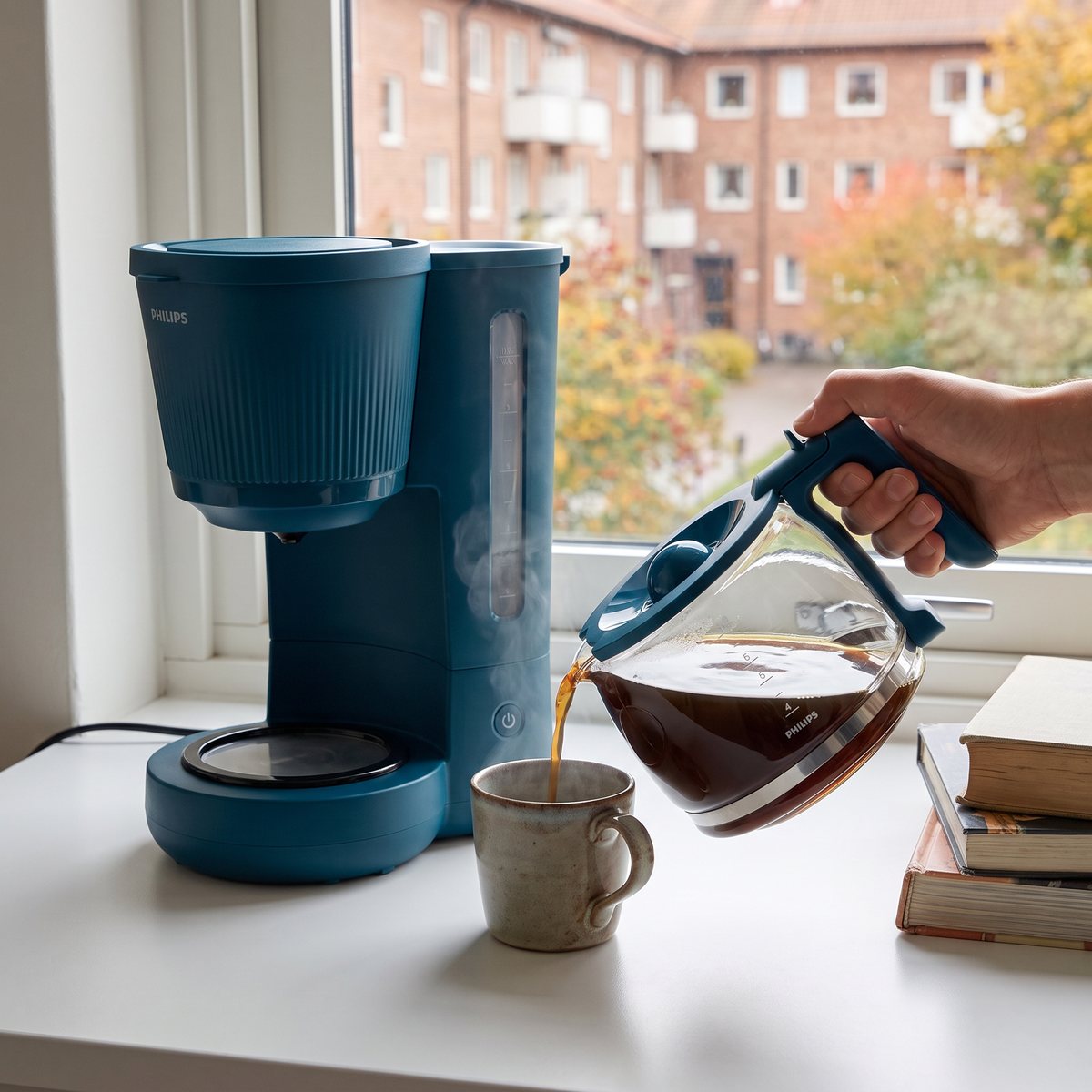 En hand häller kaffe ur glaskannan till den blå Philips-bryggaren i en mugg på en fönsterbänk i en svensk studentlägenhet med tegelhus utanför