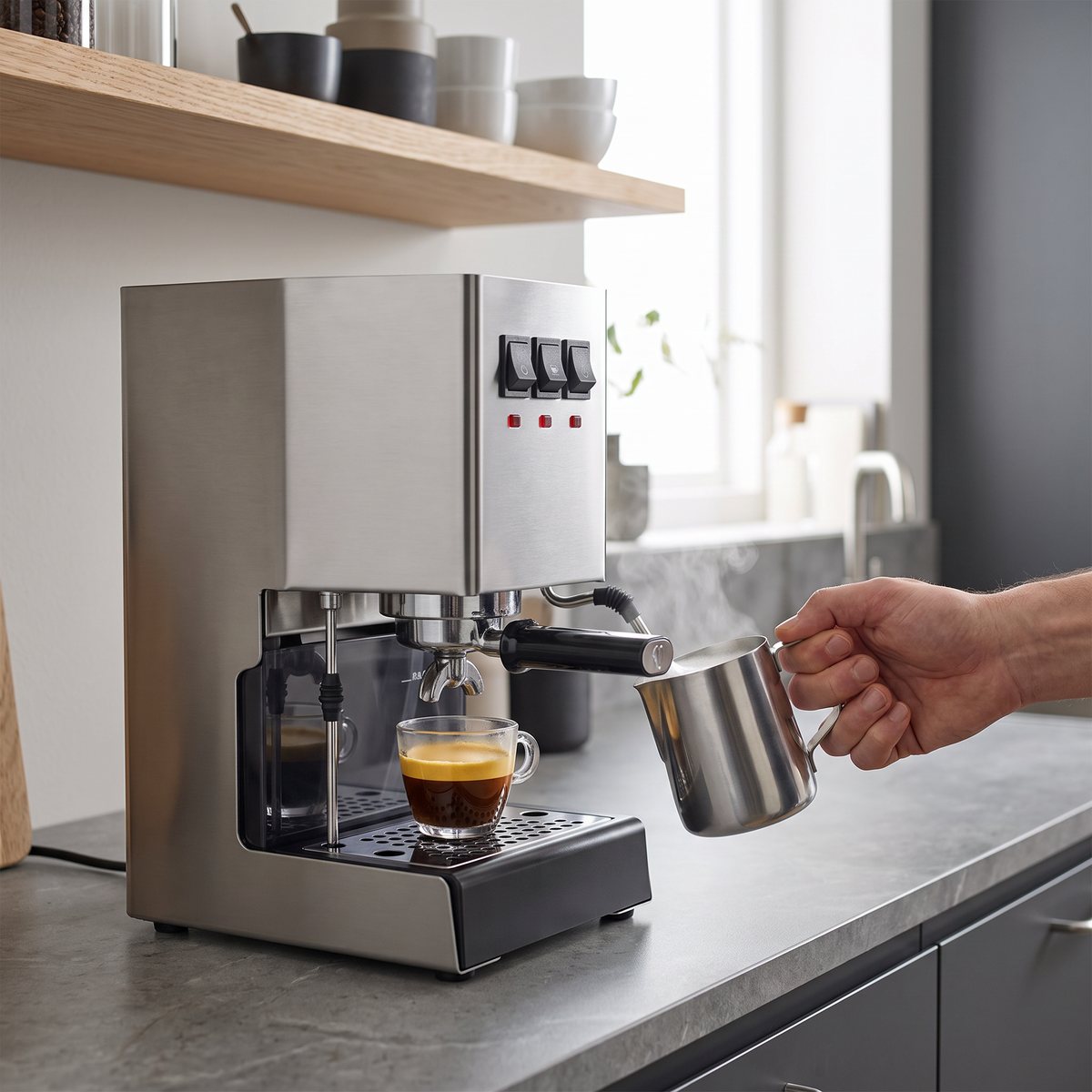 Gaggia Classic E24 i borstat stål på en grå bänkskiva i ett svenskt kök, där en hand skummar mjölk vid ångröret bredvid en kopp espresso.