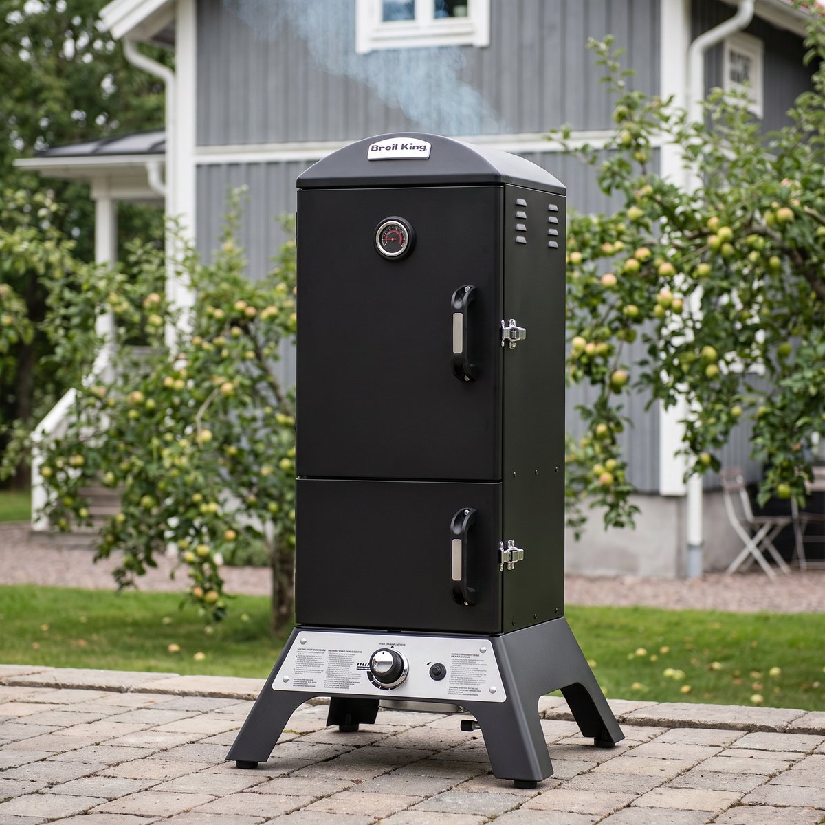 Broil King Vertical Smoker Gas på en stenlagd terrass framför ett grått trähus med äppelträd