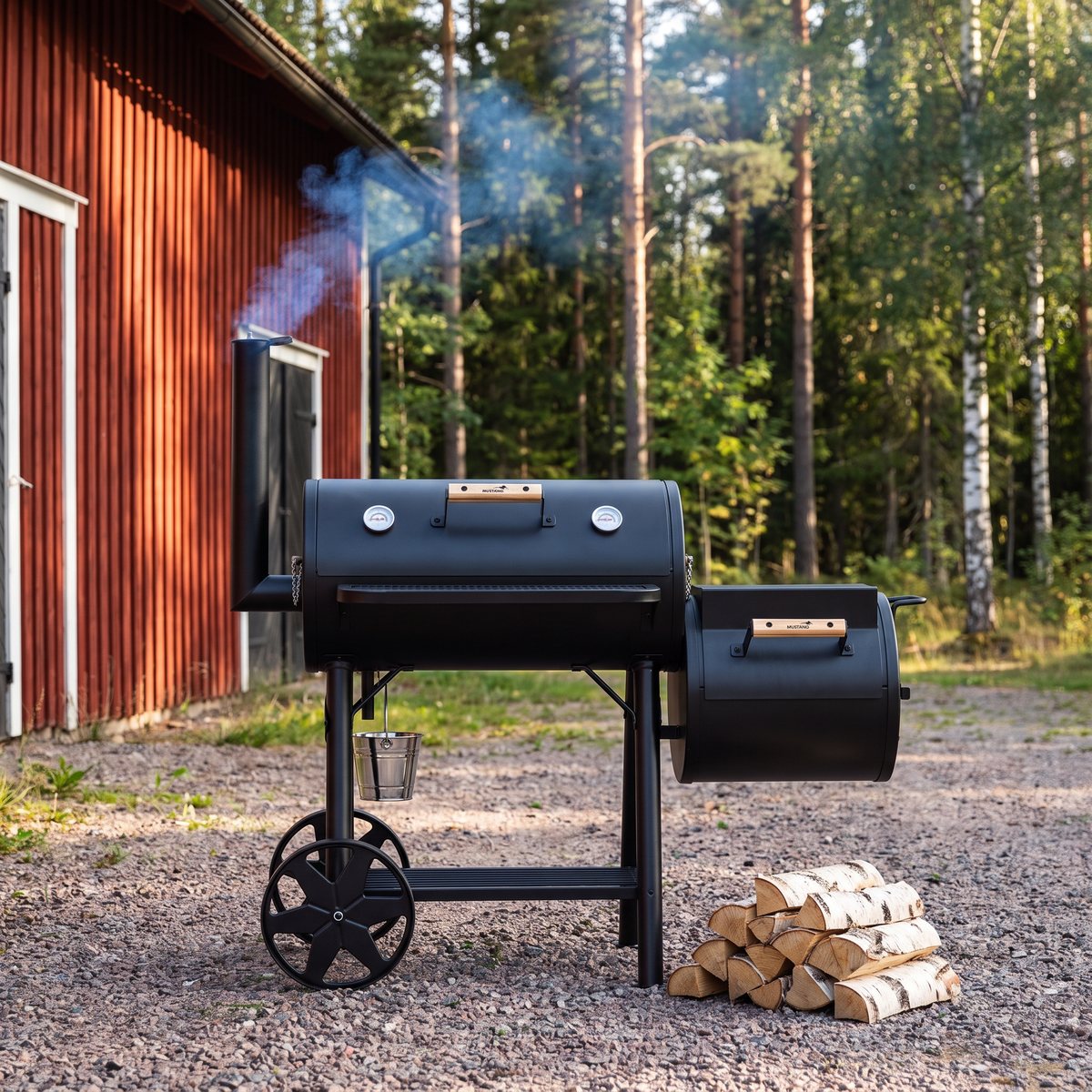 Mustang Georgia off-set smoker vid en röd lada med tall- och björkskog bakom, rök stiger ur skorstenen och björkved ligger bredvid
