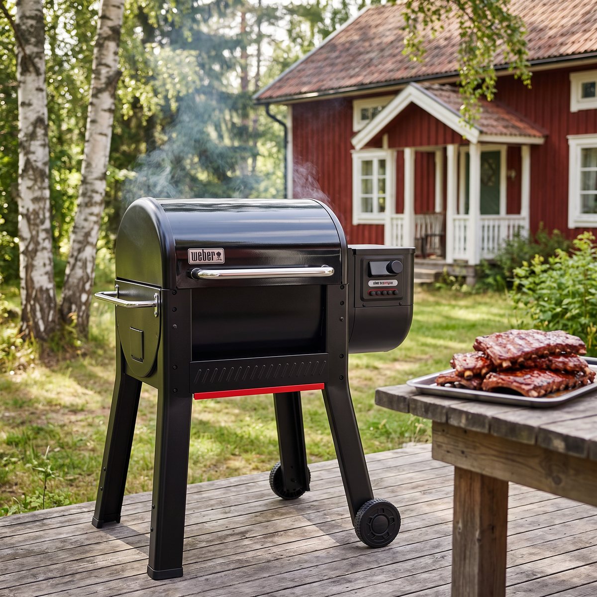Weber Smoque pelletsgrill på en trädäck vid en röd stuga med björkar, rök stiger ur grillen och en bricka med revben står på bordet