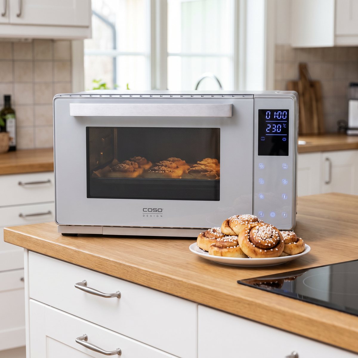 Caso Bake & Style 26 Touch i rostfritt stål på en köksbänk i ek i ett svenskt kök, med kanelbullar som gräddas och ett fat med färdiga bullar
