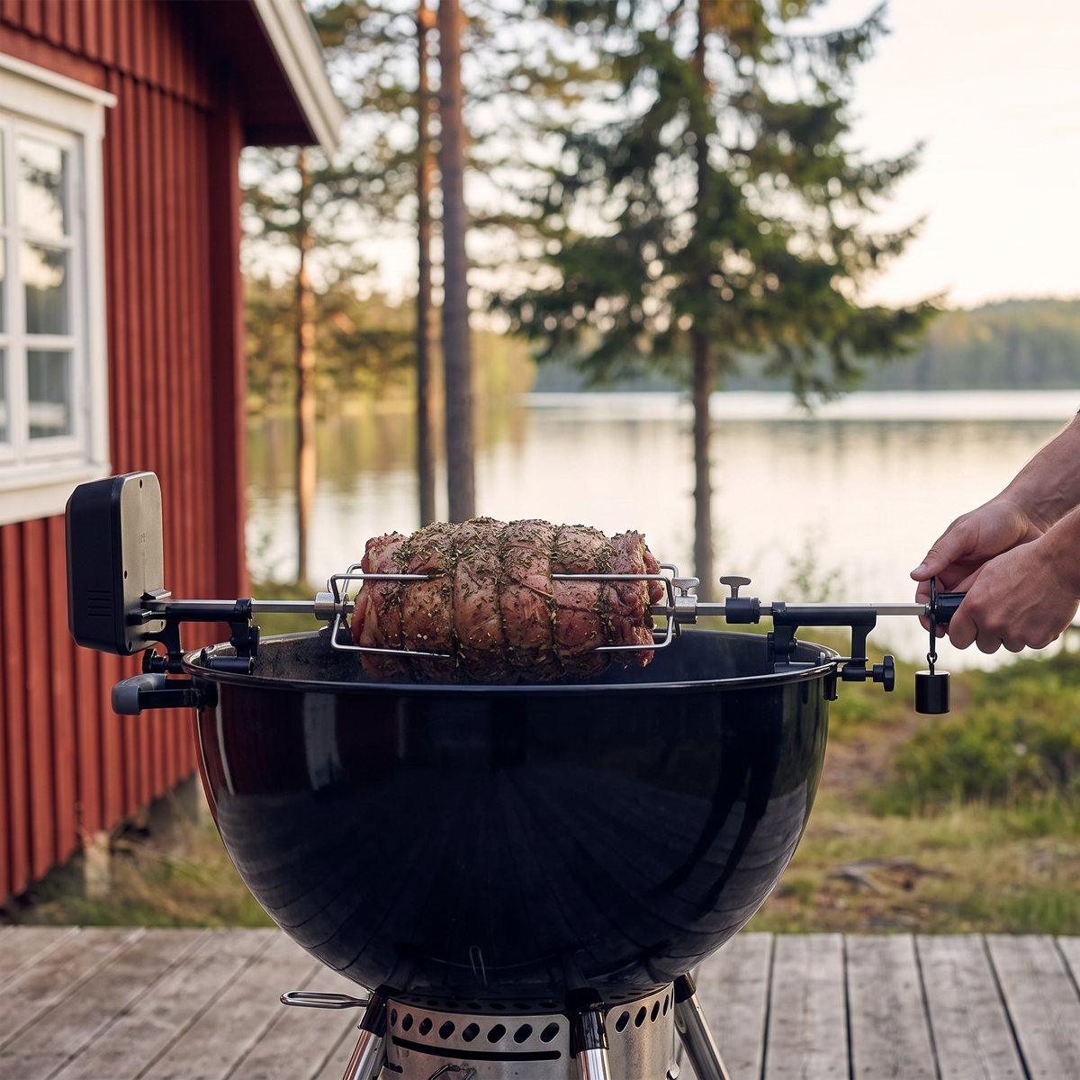 Rotisserispett med motor och klämfästen som vänder en rullstek över en svart grill på en brygga vid en svensk insjö