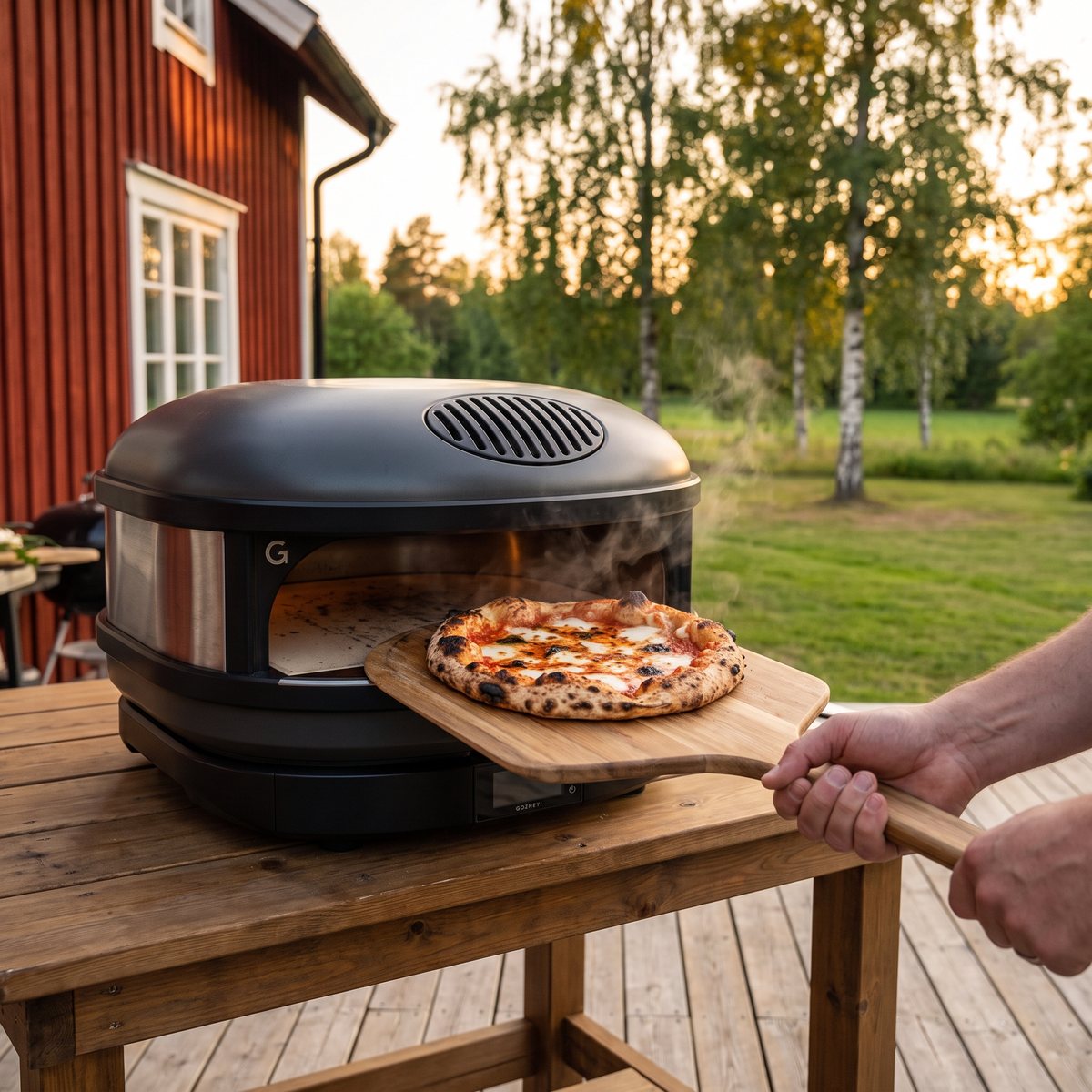 Pizzaugnen Gozney Arc på ett trädäck vid ett rött trähus, med en nygräddad pizza på pizzaspade