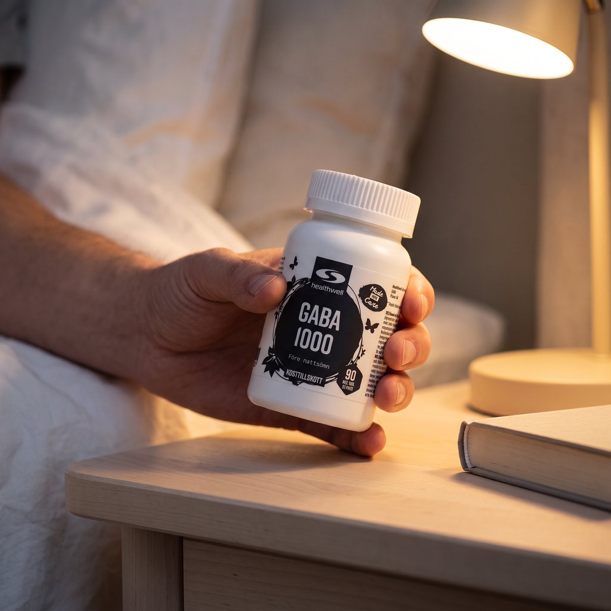 En hand håller Healthwell GABA 1000-burken över ett nattduksbord med bok och läslampa i ett svenskt sovrum