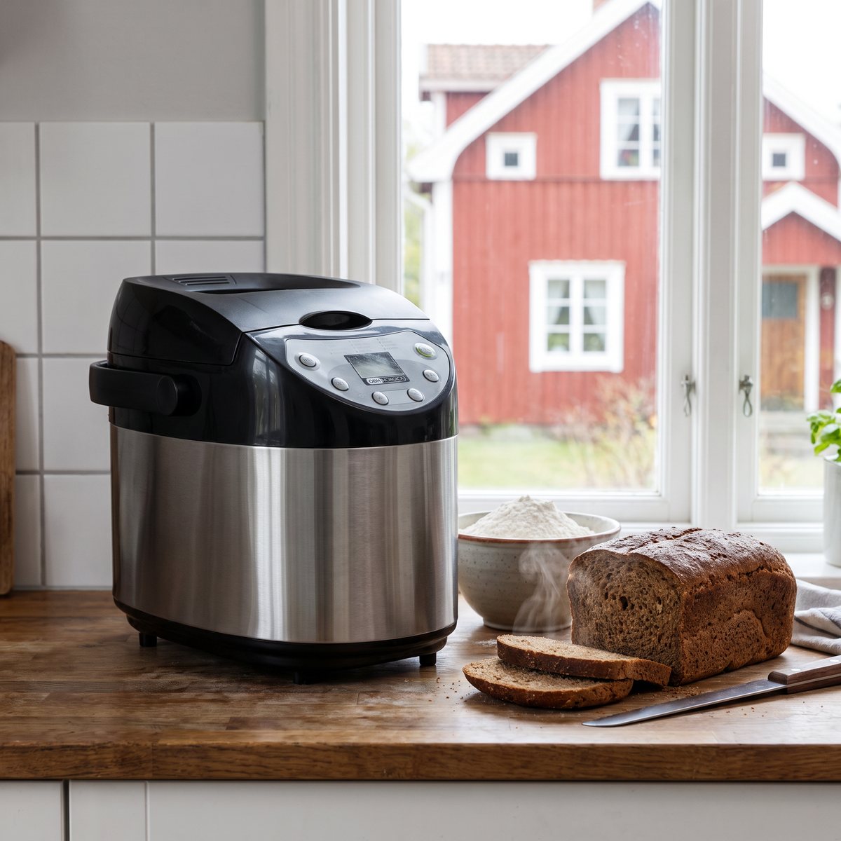 Bakmaskin i rostfritt och svart från OBH Nordica på en köksbänk med skivat mörkt bröd, med ett rött trähus synligt genom fönstret