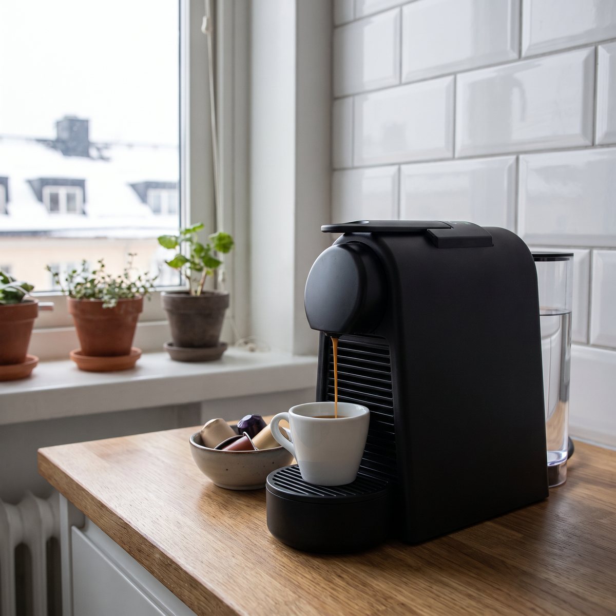Svart Nespresso Essenza Mini brygger en espresso på en smal träbänk vid fönstret i ett litet svenskt kök en vinterdag