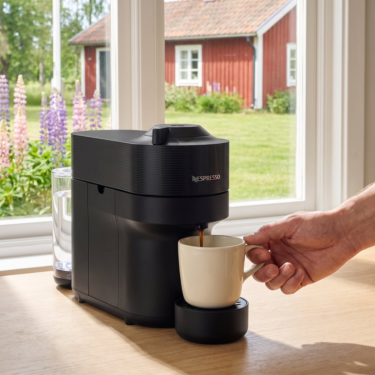Svart Nespresso Vertuo Pop fyller en mugg med kaffe på ett köksbord i en svensk sommarstuga med lupiner och rött trähus utanför