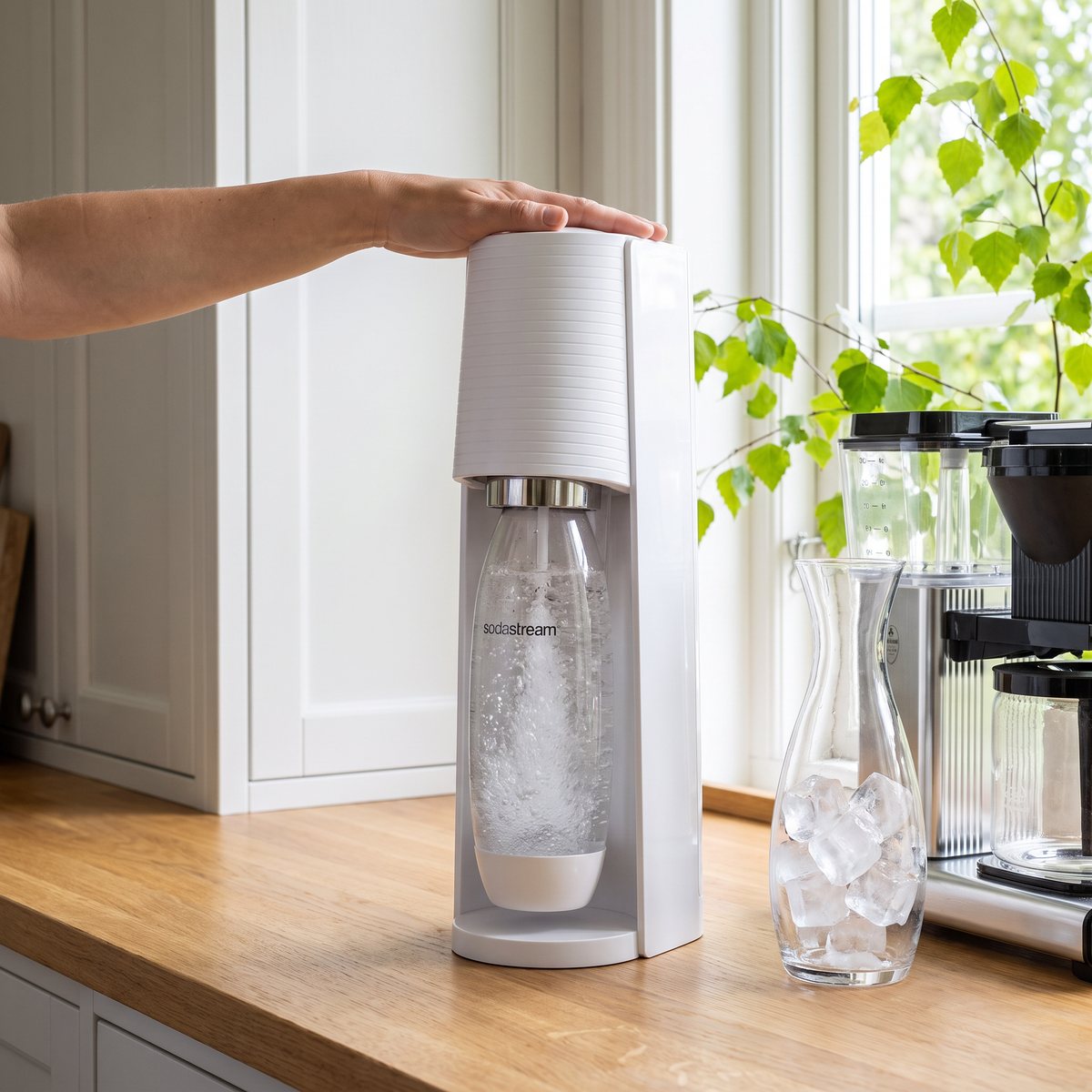 Vit Sodastream Terra där en hand trycker på kolsyreknappen, bredvid en karaff med isbitar i ett svenskt sommarkök