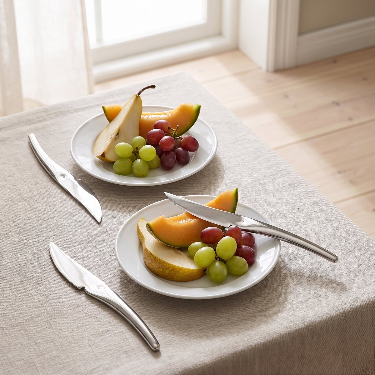 Polerade Hardanger-fruktknivar bredvid assietter med päron, melon och vindruvor på en linneduk i en ljus svensk matsal