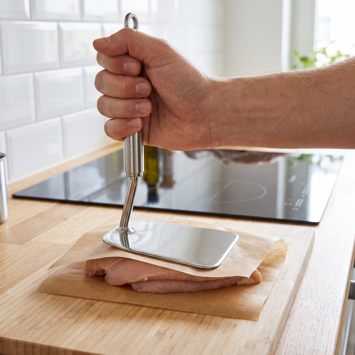Platt köttbankare i rostfritt stål plattar ut en kycklingfilé mellan bakplåtspapper i ett svenskt kök