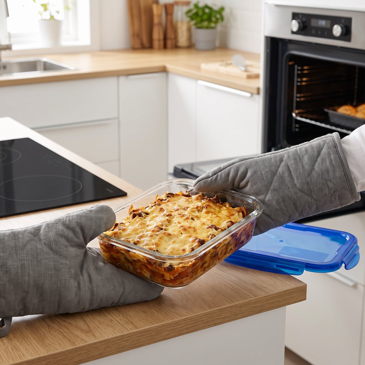 Händer i grytlappar lyfter en Pyrex Cook&Go-glaslåda med gratinerade rester ur ugnen i ett ljust svenskt kök, med det blå locket bredvid på bänken