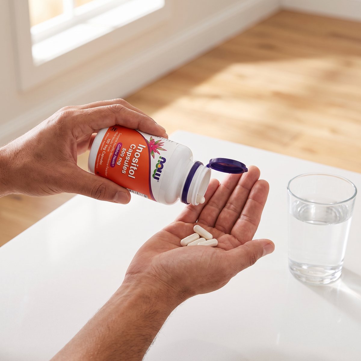 NOW Inositol 500 mg i en hand över ett glas vatten på en vit köksbänk i ett svenskt kök