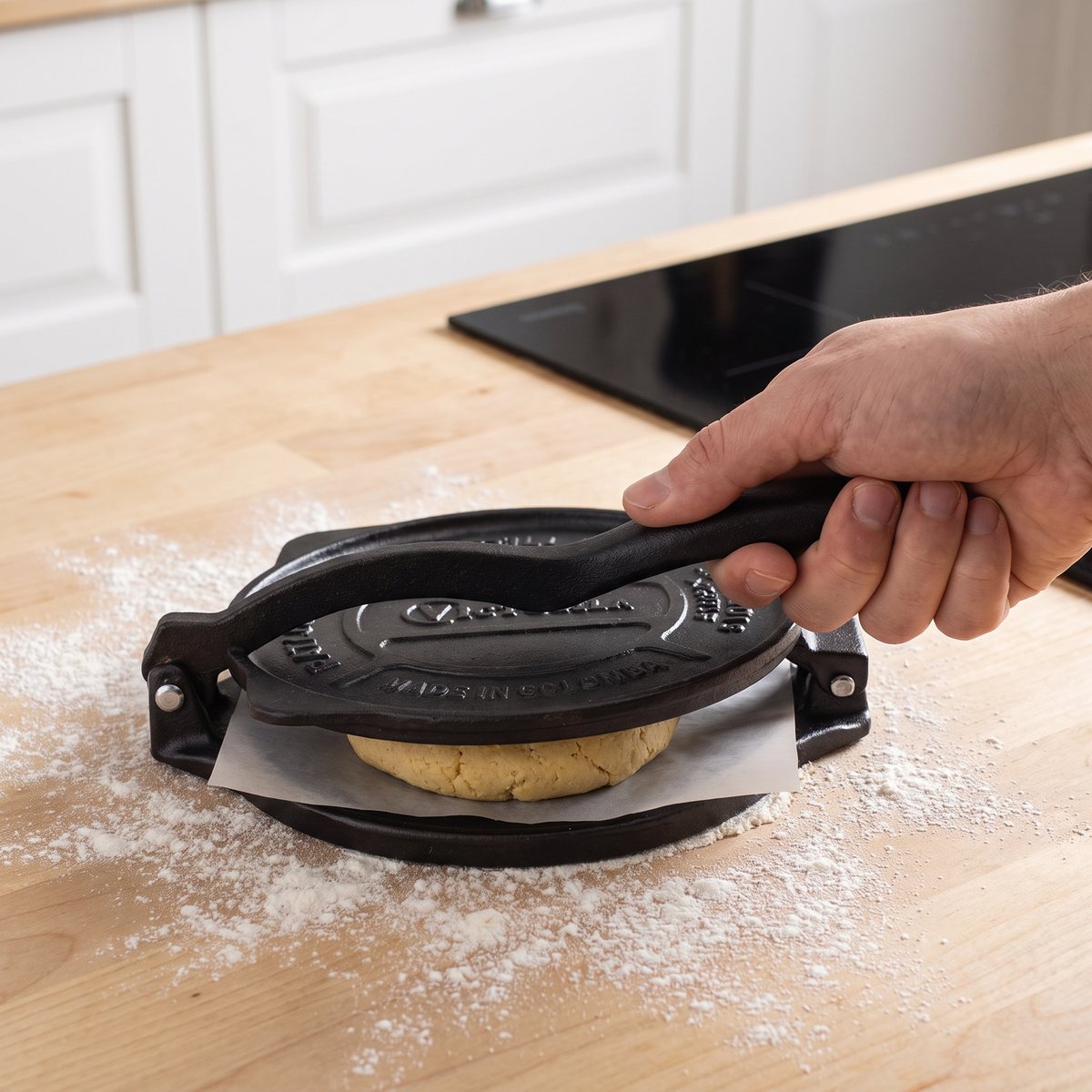 Svart tortillapress i gjutjärn pressar en degkula på en björkbänkskiva i ett svenskt kök med induktionshäll