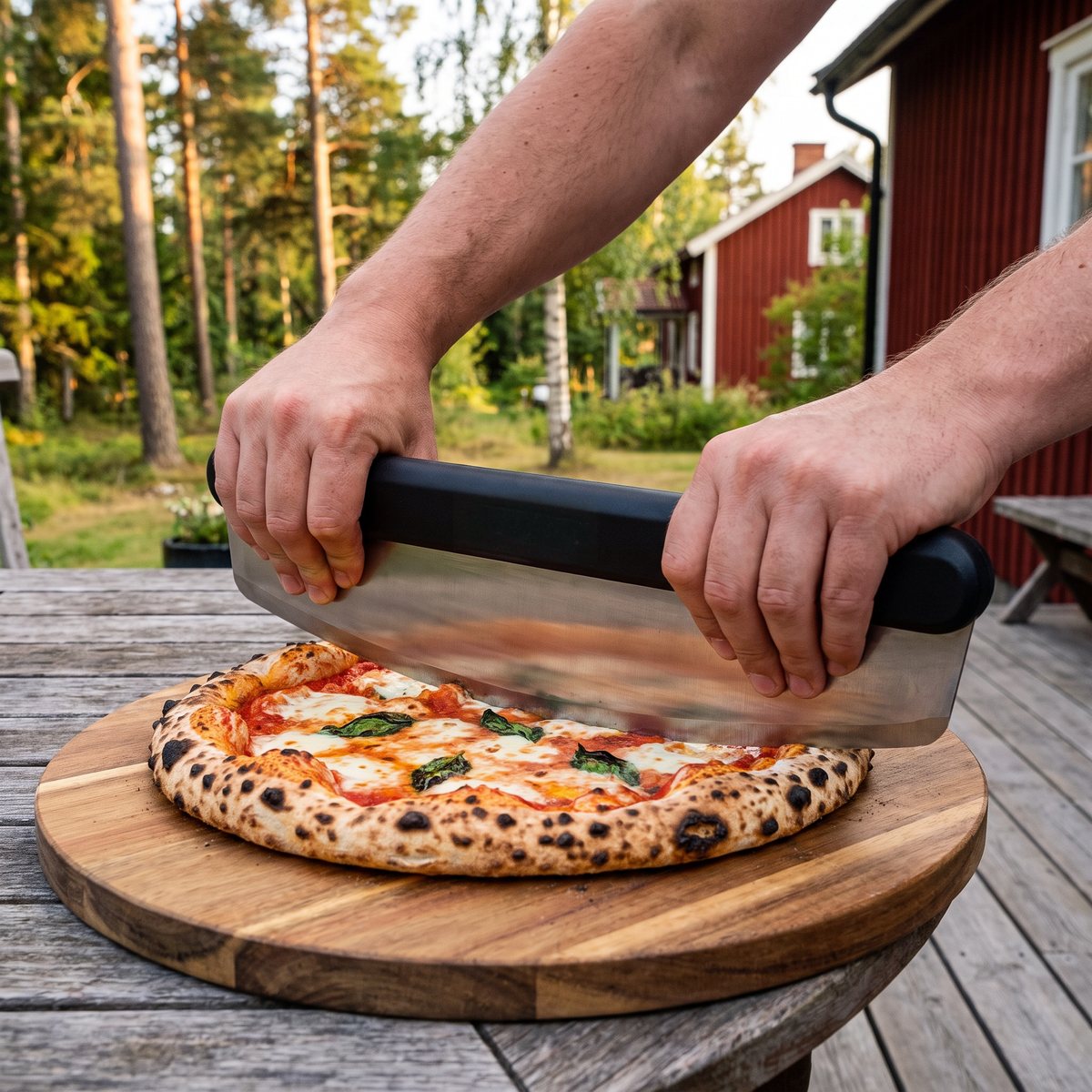 Ooni pizzaskärare vagga med svart handtag hålls med två händer över en napolitansk pizza på ett trädäck vid ett rött svenskt trähus