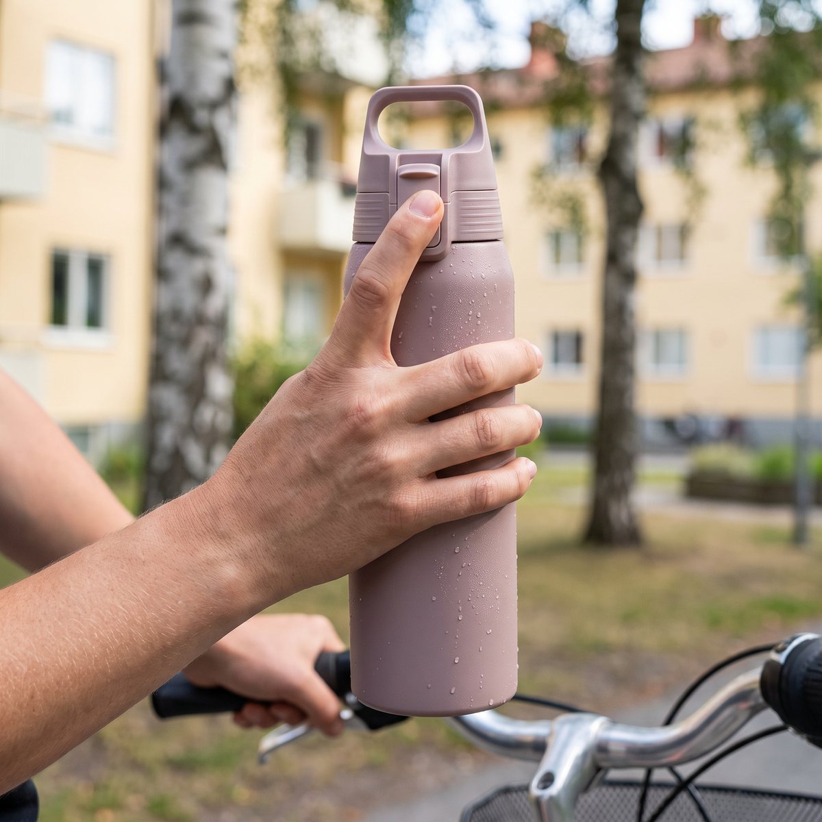 Gammalrosa Sigg Shield Therm One med kalla vattendroppar, hållen med tummen på tryckkorken vid en cykel bland björkar och bostadshus