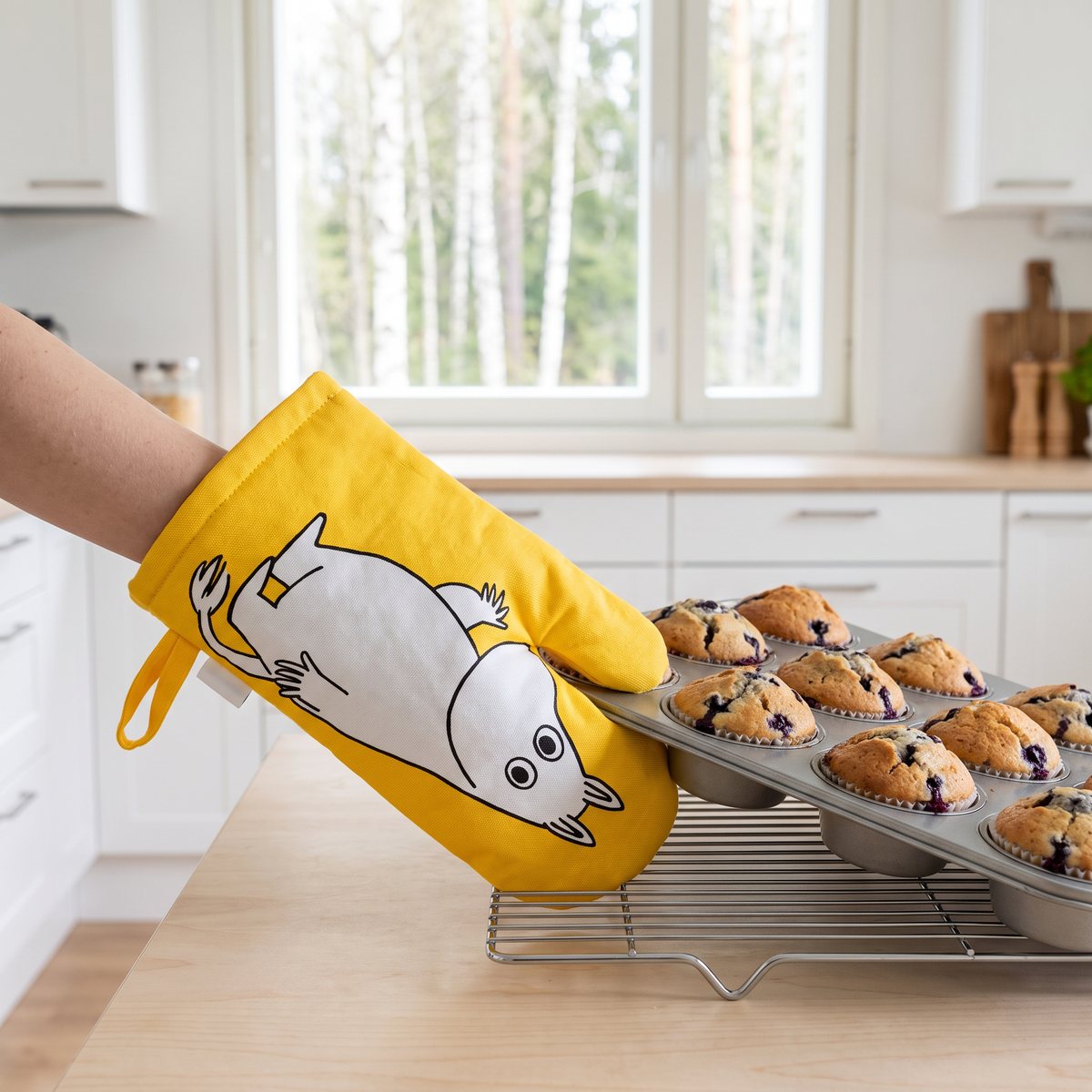 Gul Mumin-grytvante lyfter en muffinsplåt med blåbärsmuffins till ett galler i ett ljust svenskt kök med björkar utanför fönstret