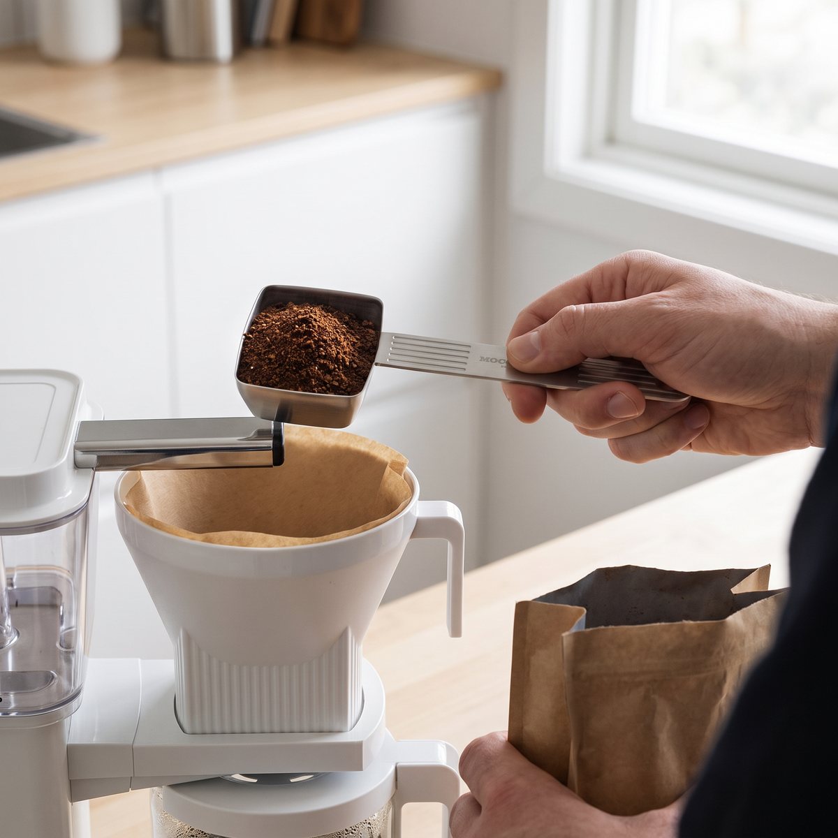 Fyrkantig måttsked i rostfritt stål med räfflat handtag, fylld med mald kaffe över ett bryggfilter i ett svenskt kök