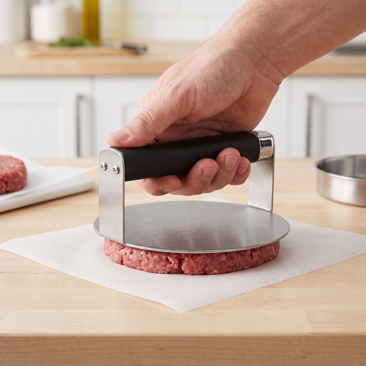 Rund burgerpress i rostfritt stål med svart handtag som pressar en köttbiff på bakplåtspapper på en träbänk i ett svenskt kök