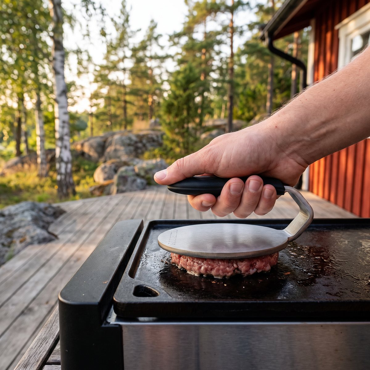 Gourmetstål Smash grillpress pressar en burgare på en stekhäll på en trädäck vid en röd stuga i svensk natur