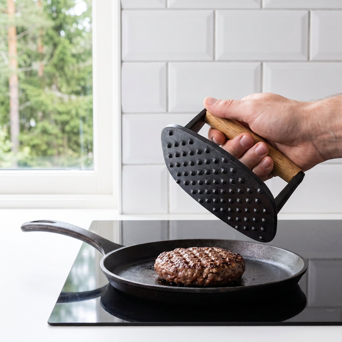 Ronneby Bruk Maestro burgerpress med knottrig undersida lyfts från en burgare i en gjutjärnspanna på induktionshäll i ett svenskt kök