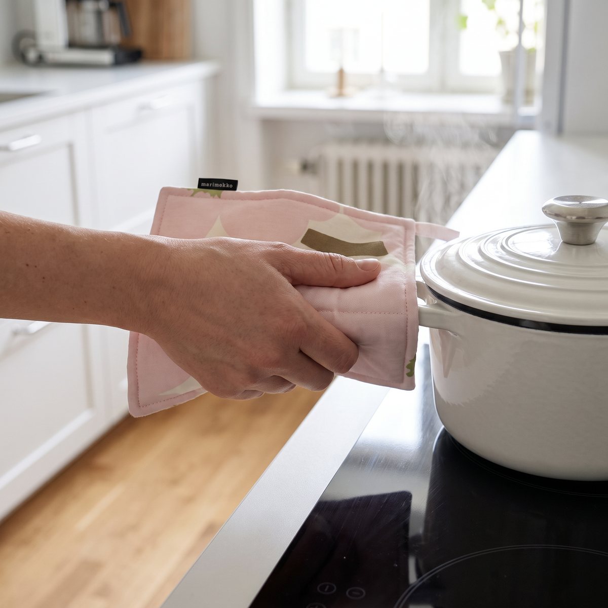 Rosa grytlapp från Marimekko med Unikko-mönster som en hand håller runt handtaget på en het gryta på en induktionshäll i ett svenskt kök.