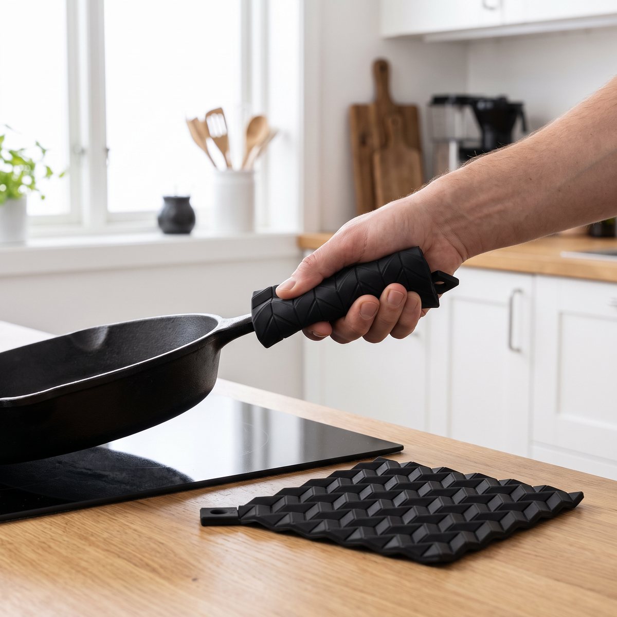 Två svarta silikongrytlappar med relief från RIG-TIG, där en hand lyfter en het gjutjärnspanna från en induktionshäll i ett svenskt kök.