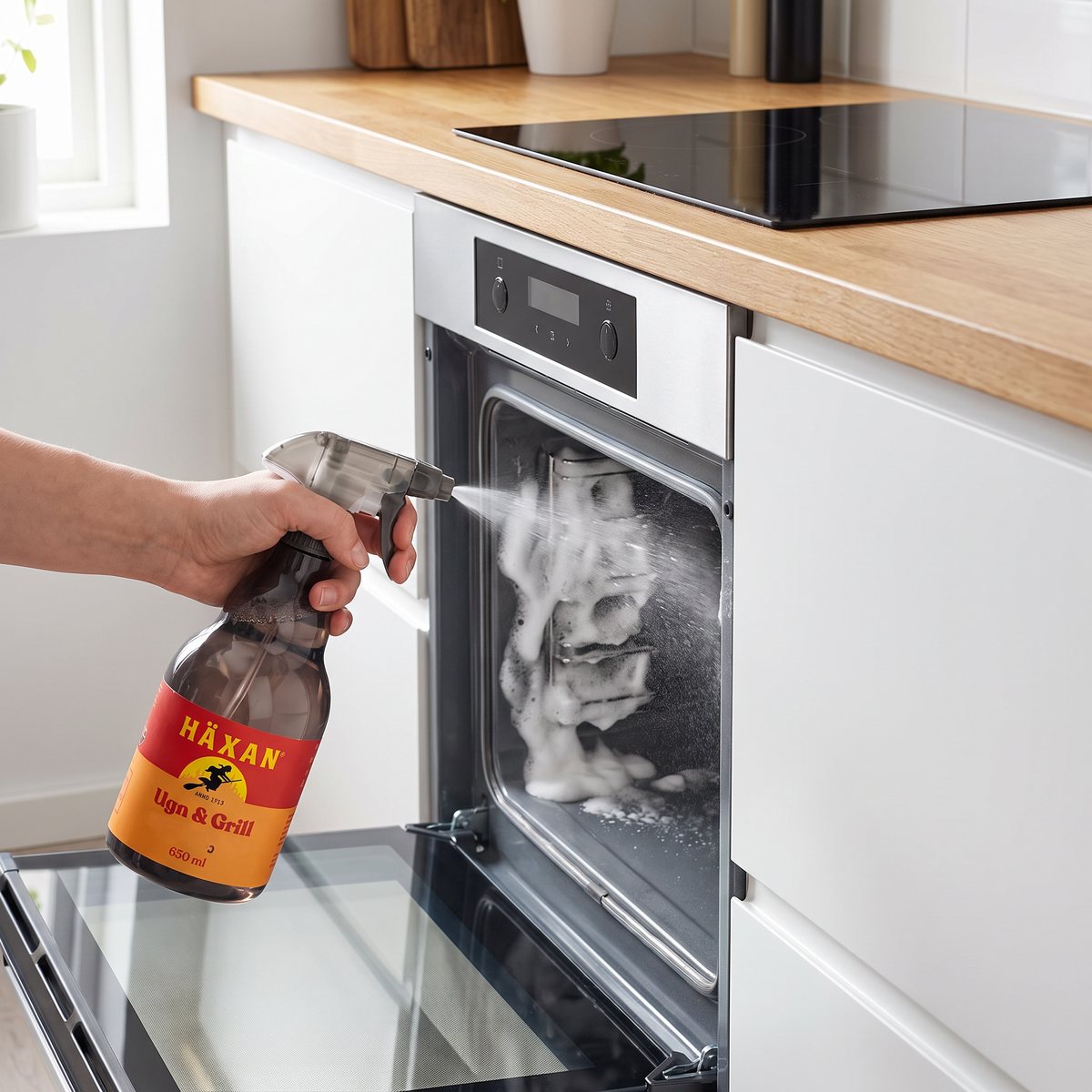 Hand sprayar Häxan Ugn & Grill så att skummet lägger sig på väggen i en öppen inbyggd ugn under en induktionshäll i ett ljust svenskt kök