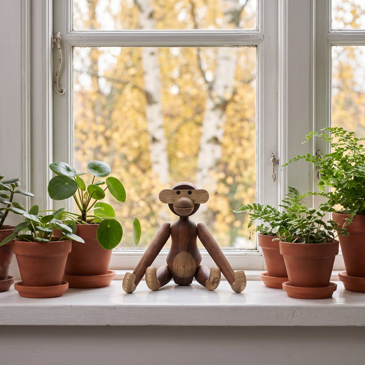 Kay Bojesen Apa Mini i teak sitter på en fönsterbräda mellan krukväxter, med björkar i höstfärger utanför fönstret