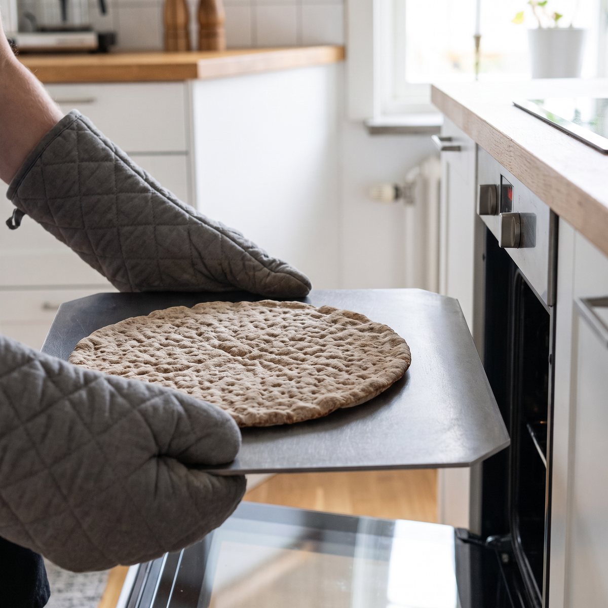 Gourmetstål Light, en grå stålplatta med avfasade hörn, lyfts ur ugnen med grytlappar i ett ljust svenskt kök
