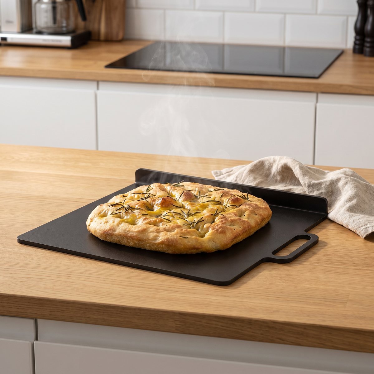 Kockums Jernverk bak- och pizzastål i svart kolstål med uppvikt kant och handtag, med en rosmarinfocaccia på en bänkskiva i ek framför en induktionshäll i ett svenskt kök