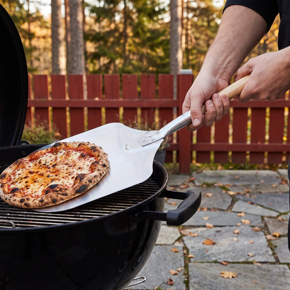 Kitchencraft pizzaspade med trähandtag som lyfter en pizza från grillgallret på en kolgrill på en stenlagd uteplats