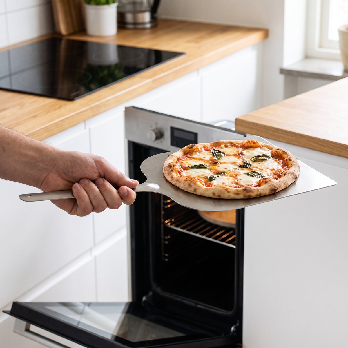 Kockums Jernverk pizzaspade i rostfritt stål med en gräddad pizza framför en inbyggd ugn i ett ljust svenskt kök