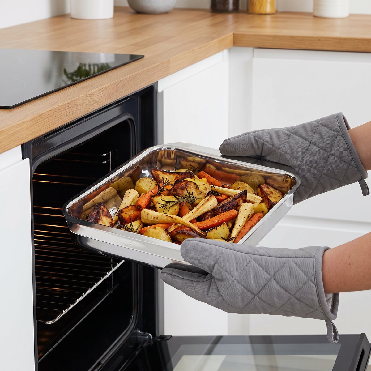 Tefal Excellence ugnsform i rostfritt stål med rostade rotfrukter lyfts ut ur ugnen med grytlappar i ett vitt svenskt kök med induktionshäll
