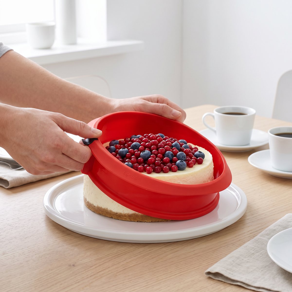 Lékué springform där den röda silikonringen viks undan från en cheesecake med bär på det vita keramikfatet, på ett svenskt fikabord