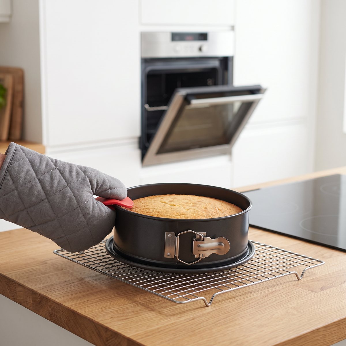 Woll Bake It springform med nygräddad sockerkaka hålls i det röda silikongreppet med grytvante på ett galler, i ett svenskt kök med induktionshäll