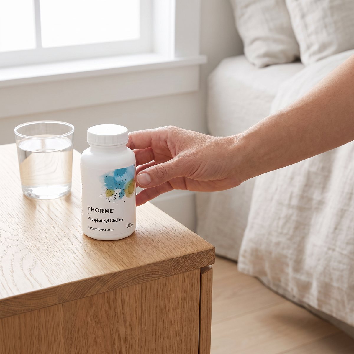 Thorne Phosphatidyl Choline på ett nattduksbord i ett svenskt sovrum, med ett glas vatten och en hand som greppar burken