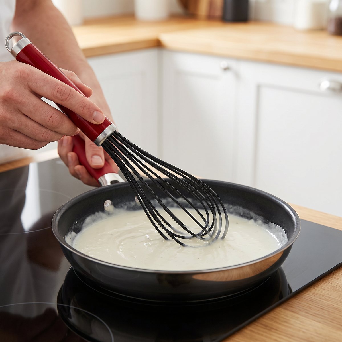 KitchenAid ballongvisp med röd handtag och silikontrådar som rör i vit sås i en stekpanna på en induktionshäll i ett svenskt kök