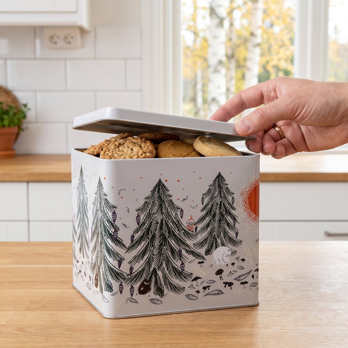 Avlång grå plåtburk med Mumin-skogsmotiv där locket lyfts över kakor på en köksbänk i ett svenskt kök