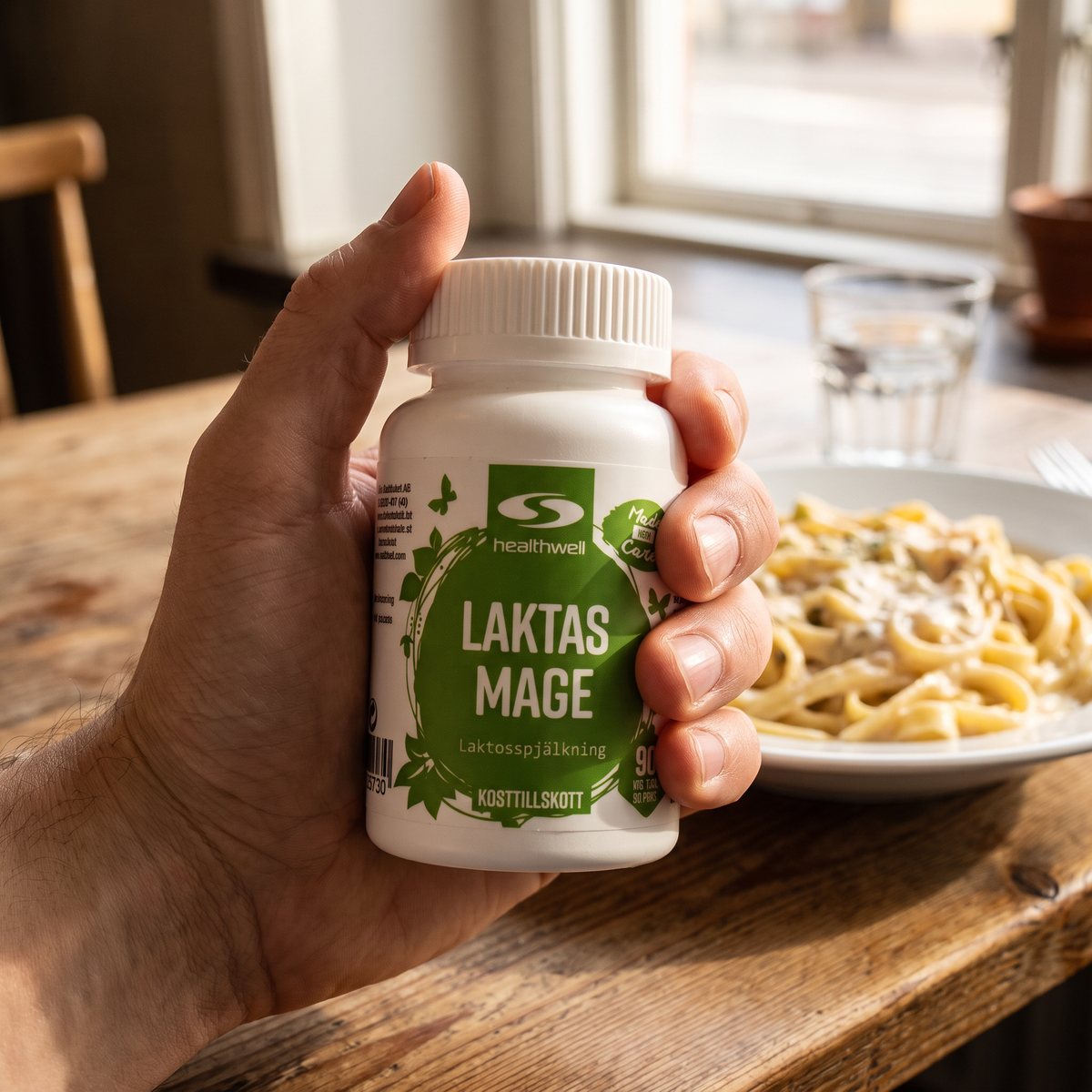 Hand som håller en burk Healthwell Laktas Mage över ett cafébord med pasta i en svensk miljö