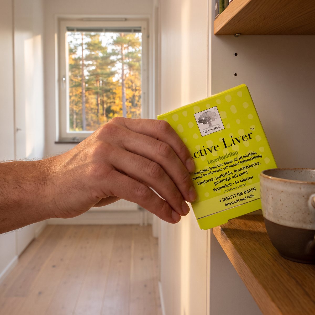 Hand som tar New Nordic Active Liver från en hylla i ett svenskt hem med tallskog utanför fönstret