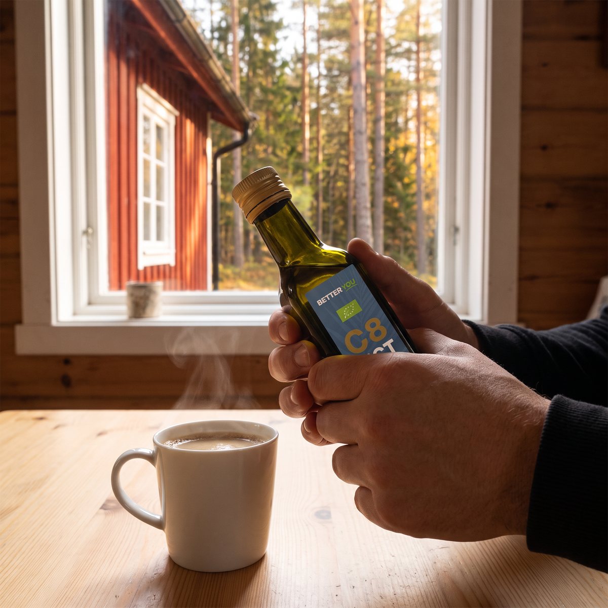 Better You C8 MCT-olja i liten glasflaska hållen över en kaffekopp i en svensk stuga