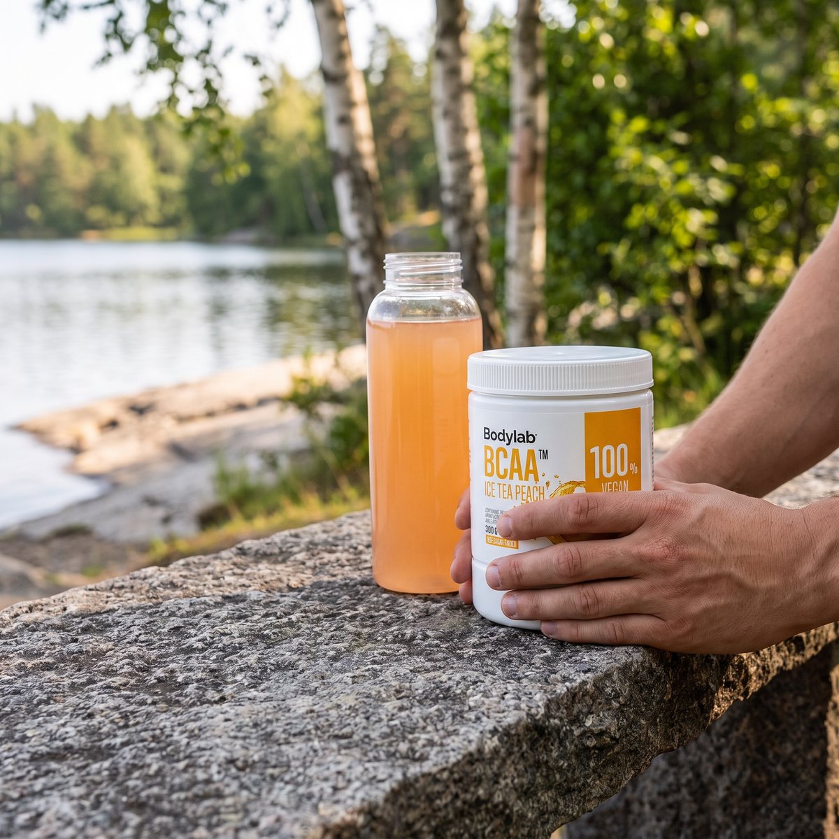 Burk med Bodylab BCAA på en granitbänk vid en svensk sjö med björkar och en flaska med persikodryck