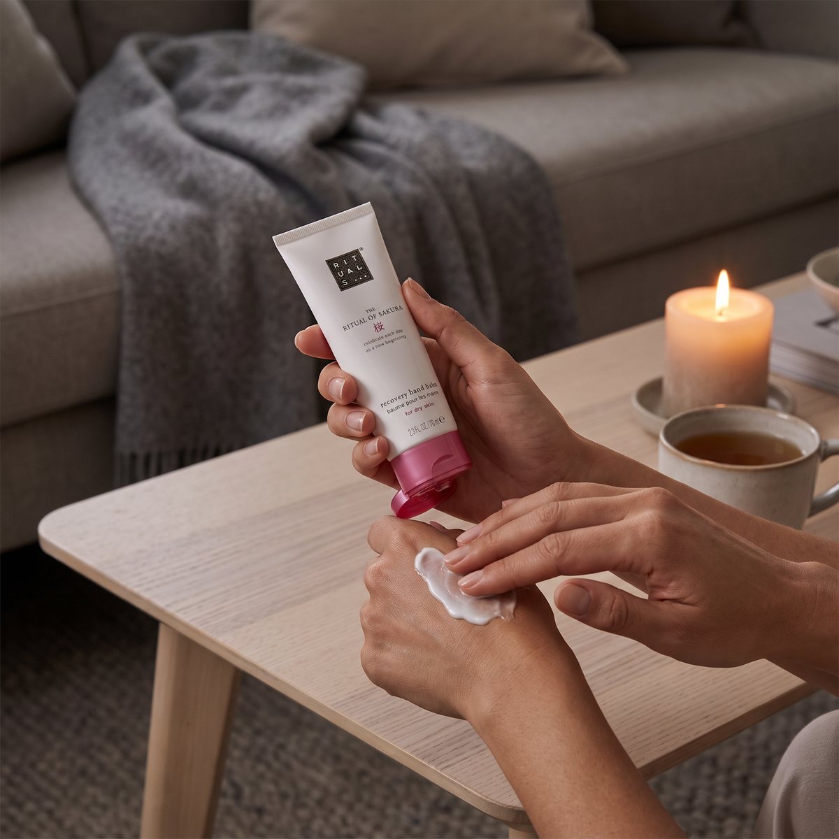 Rituals Sakura Recovery Hand Balm som masseras in i huden i en soffa i ett mysigt svenskt vardagsrum