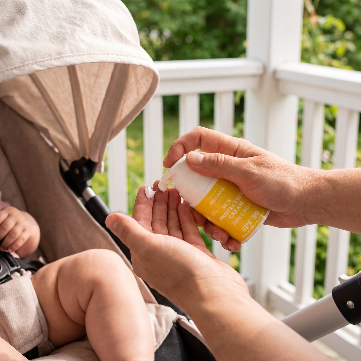 Föllinge Sun Protection Baby & Kids pumpas ut på fingertopparna bredvid en bebis i barnvagn på en vit veranda