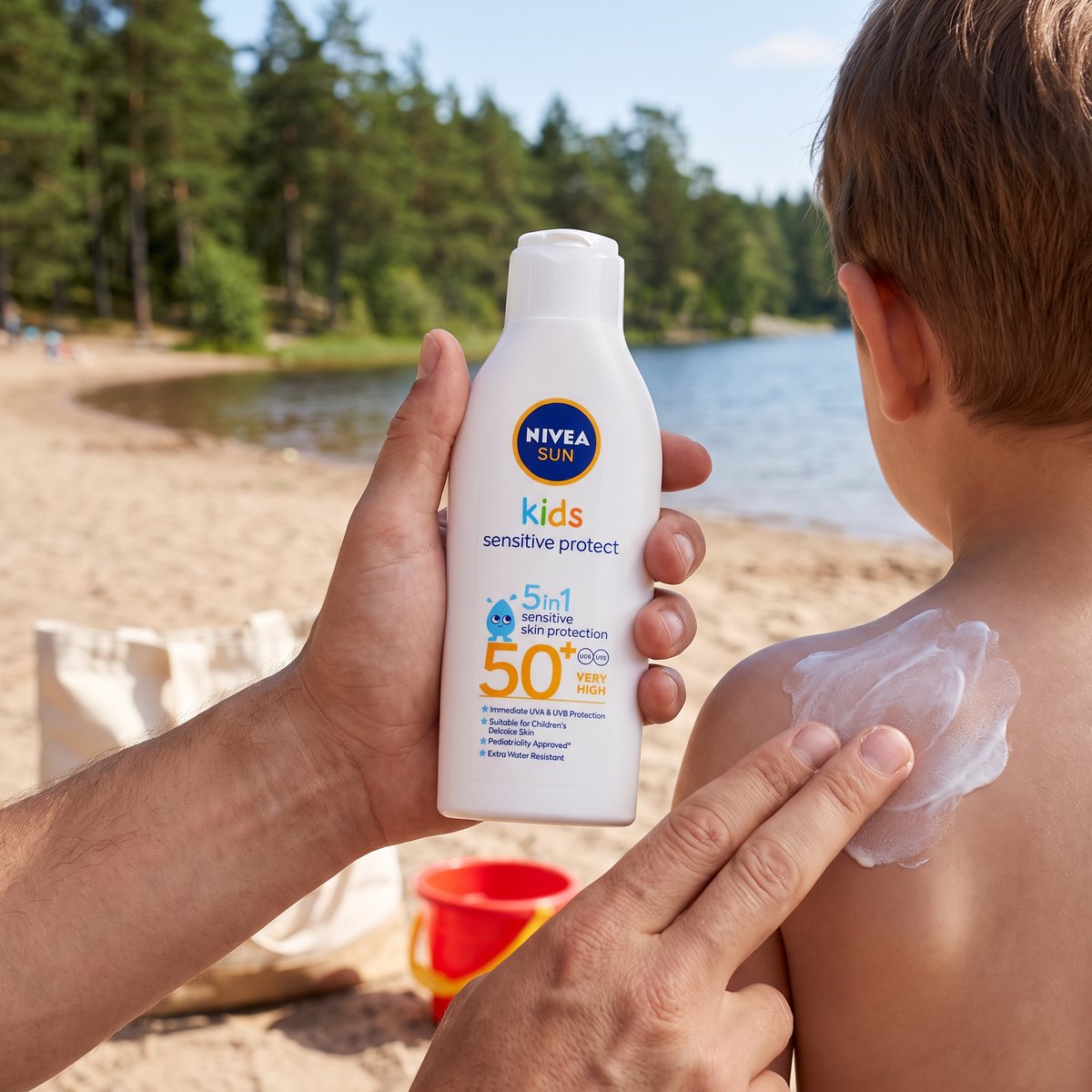 NIVEA Sun Kids Sensitive Protect & Play hålls upp medan lotionen smörjs på ett barns axel på en sandstrand vid en svensk skogssjö