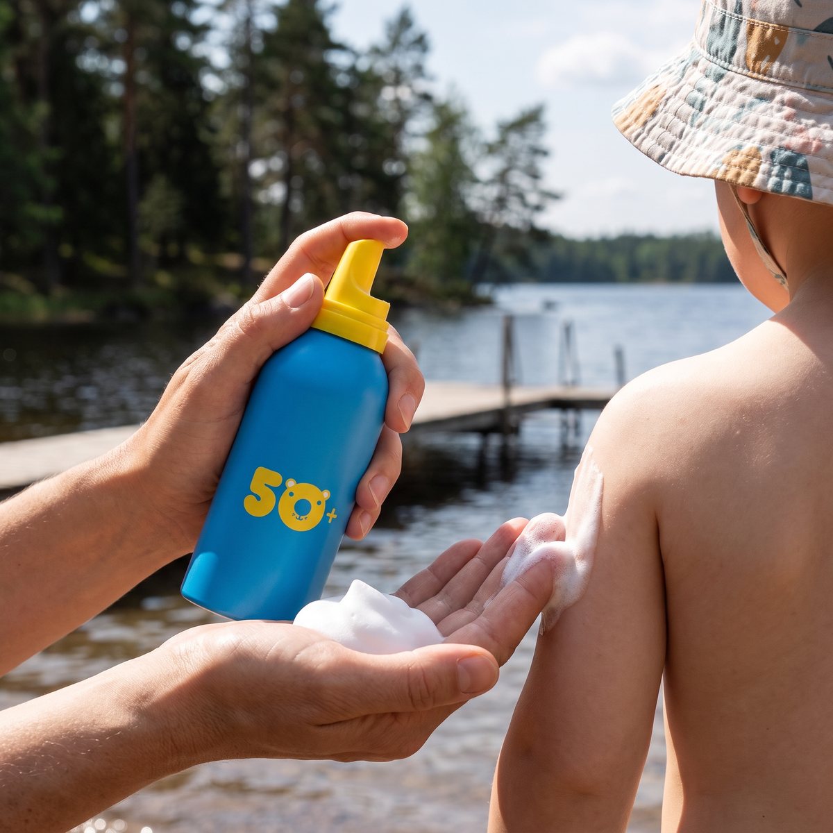 EVY Kids SPF 50 trycks ut i en vuxens handflata och smörjs på ett barns axel vid en badbrygga i en svensk insjö