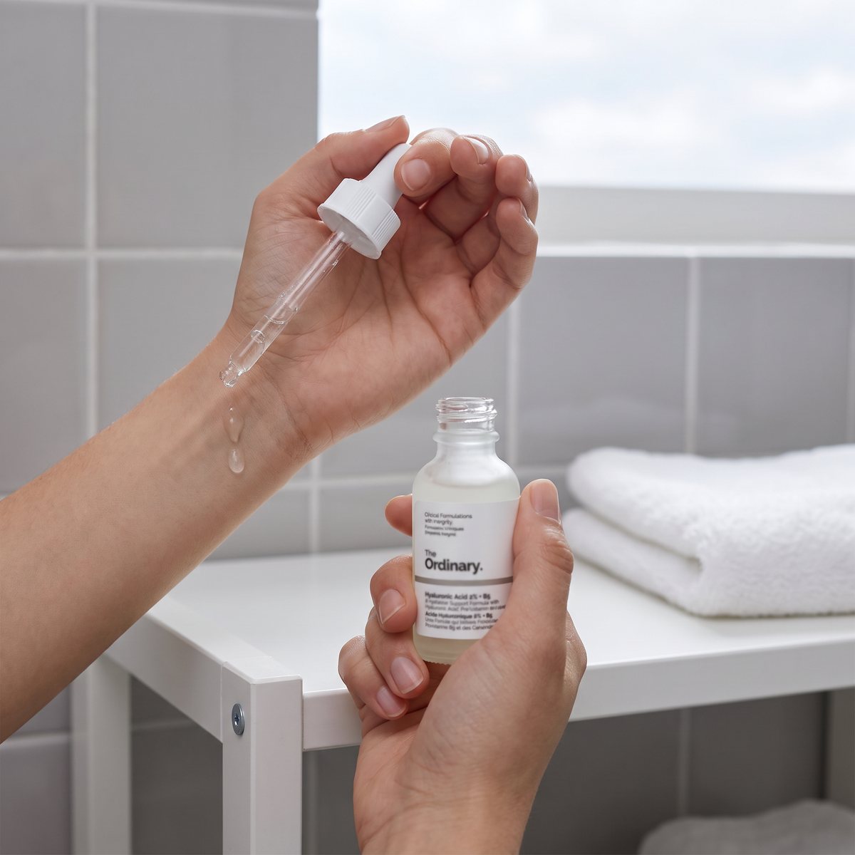 Händer som droppar The Ordinary Hyaluronic Acid 2% + B5 på handleden vid en vit badrumshylla i en svensk lägenhet