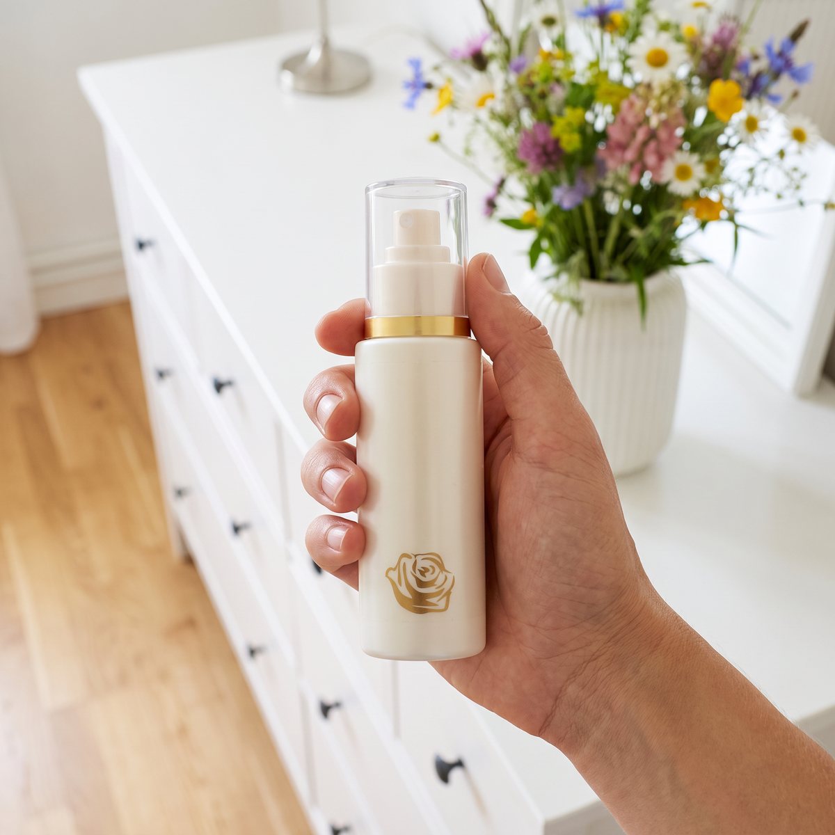 Rosenserien deodorantspray hålls i handen vid en vit byrå i ett ljust svenskt sovrum med vilda blommor